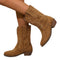 Bottes cowboy en daim marron portées par une femme, présentées en gros plan sur un sol en bois texturé. Le modèle montre des broderies décoratives et un talon bas, créant un style western tendance et décontracté.