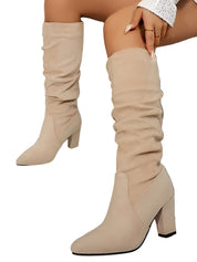 Bottes hautes en daim beige portées par une femme assise, présentées en gros plan dans un intérieur moderne. Le modèle possède un talon épais, une tige légèrement plissée et une finition élégante qui met en valeur un style contemporain et raffiné.