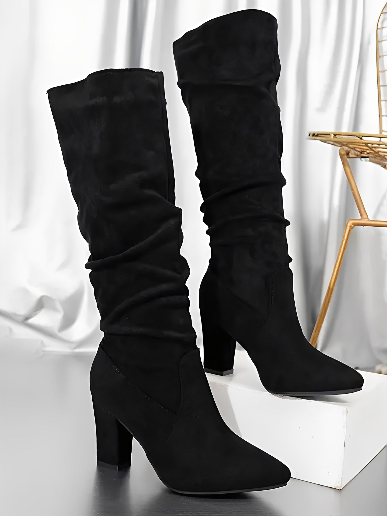 Paire de bottes hautes en daim noir présentées sur une surface lumineuse, avec un talon épais élégant et une tige légèrement plissée. Les bottes affichent une silhouette moderne et raffinée, mises en valeur dans un décor intérieur aux tons métallisés.