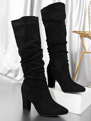 Paire de bottes hautes en daim noir présentées sur une surface lumineuse, avec un talon épais élégant et une tige légèrement plissée. Les bottes affichent une silhouette moderne et raffinée, mises en valeur dans un décor intérieur aux tons métallisés.