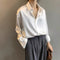 Une femme porte une chemise blanche en satin, légèrement boutonnée, avec un pantalon gris à plis. Elle porte également un collier simple en or et tient un sac à main marron dans ses mains, ajoutant une touche élégante à son look décontracté.