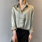 Une femme porte une chemise satinée de couleur vert olive avec un col classique et des manches retroussées. Le haut est boutonné, et elle porte un pantalon noir à taille haute. Elle ajoute une touche élégante avec un collier délicat en or et un sac marron. Son look est raffiné et moderne.