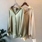 Deux chemises en satin élégantes accrochées sur des cintres. L'une est de couleur vert olive, tandis que l'autre est de couleur beige clair. Les deux chemises présentent un col classique et des manches longues, avec des poignets légèrement retroussés.