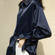Une chemise en satin bleu nuit portée par une femme. Elle a un col classique, des manches longues légèrement retroussées, et la coupe fluide crée une allure élégante et décontractée.