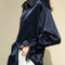 Une chemise en satin bleu nuit portée par une femme. Elle a un col classique, des manches longues légèrement retroussées, et la coupe fluide crée une allure élégante et décontractée.