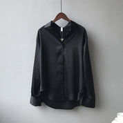 Une chemise en satin noir élégante accrochée à un cintre. Elle possède un col classique, des manches longues avec des poignets boutonnés et une coupe fluide qui lui confère un style moderne et sophistiqué.