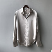 Une chemise en satin beige élégante accrochée à un cintre. Elle présente un col classique, des manches longues avec des poignets boutonnés, et une coupe fluide qui lui donne un aspect raffiné et moderne.