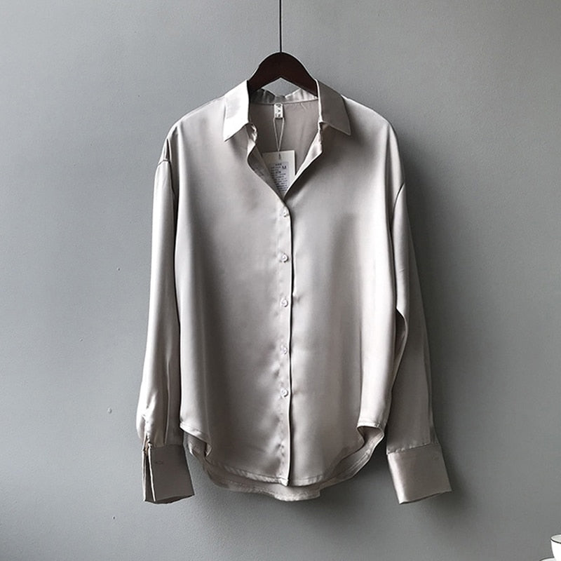 Une chemise en satin beige élégante accrochée à un cintre. Elle présente un col classique, des manches longues avec des poignets boutonnés, et une coupe fluide qui lui donne un aspect raffiné et moderne.